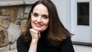 Woman in black turtle-neck shirt with brown hair, leaning chin on right fist, smiling