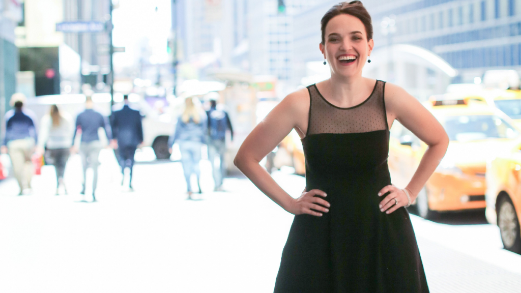 Smiling woman with hands on her hips stands on bustling street with yellow taxi cabs in the background, text reads: Our one hundredth episode, fan favorites, editor's pick and the best clips and highlights.