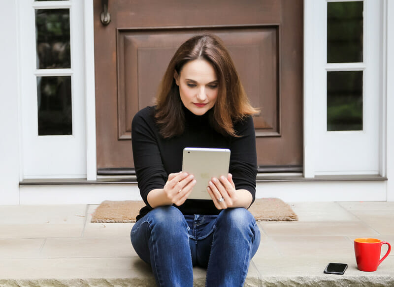 A woman sits on steps staring intently at her tablet. Her phone and red coffee mug are beside her.