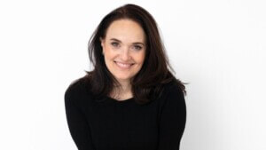 photo of woman with dark brown, shoulder-length hair in front of a white wall smiling, wearing a black long-sleeve shirt
