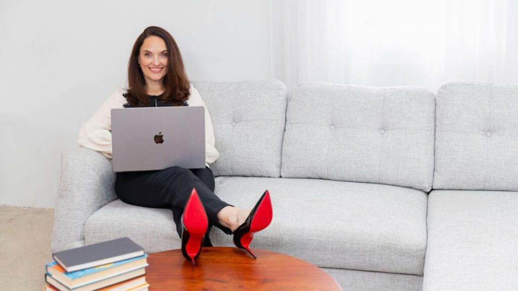 Sarah Walton wearing white blouse and black slacks sitting on a light gray sofa with laptop on her lap