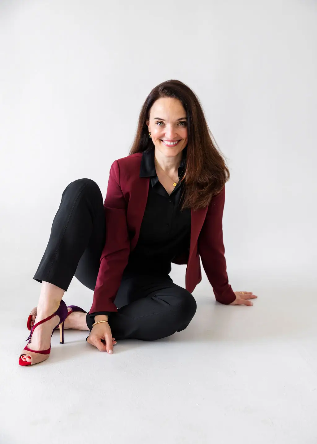 Sarah Walton wearing burgundy blazer sitting on white floor on a white background