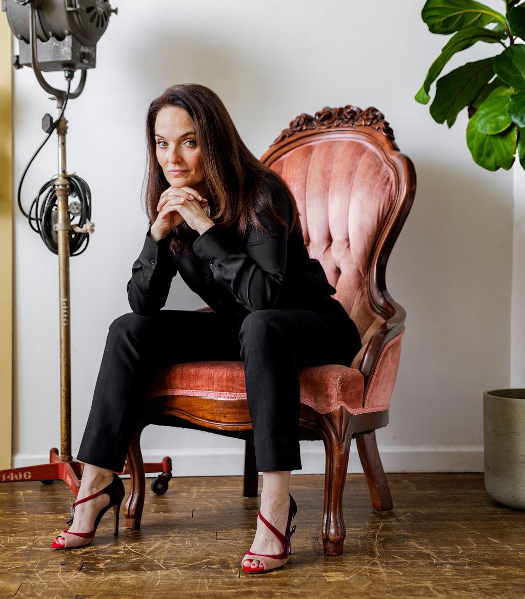 Sarah Walton sitting in an ornate pink chair wearing a black outfit looking serious