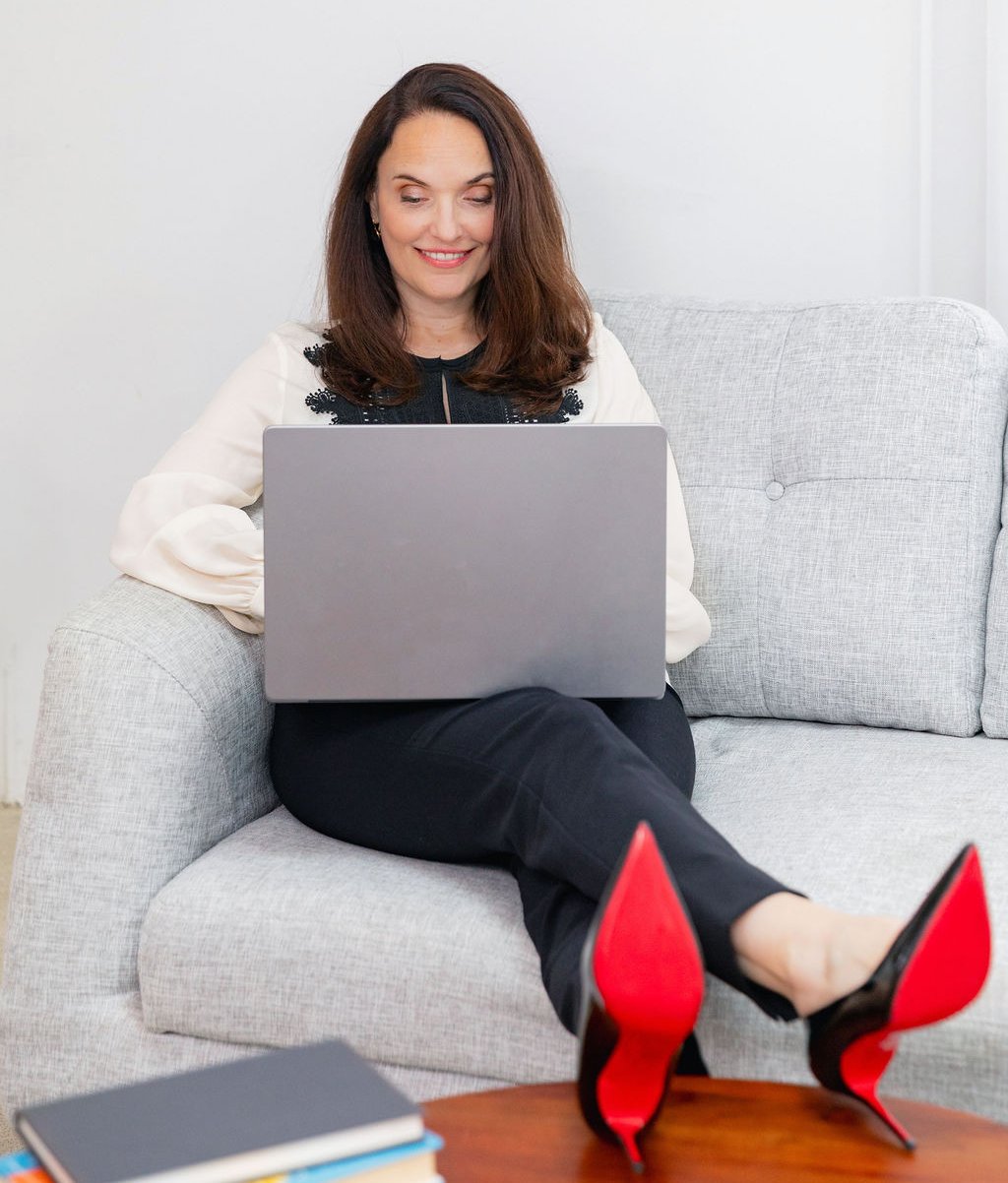 Sarah Walton seated on a couch working on a laptop, wearing red bottomed heels.