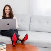 Sarah Walton wearing white blouse and black slacks sitting on a light gray sofa with laptop on her lap