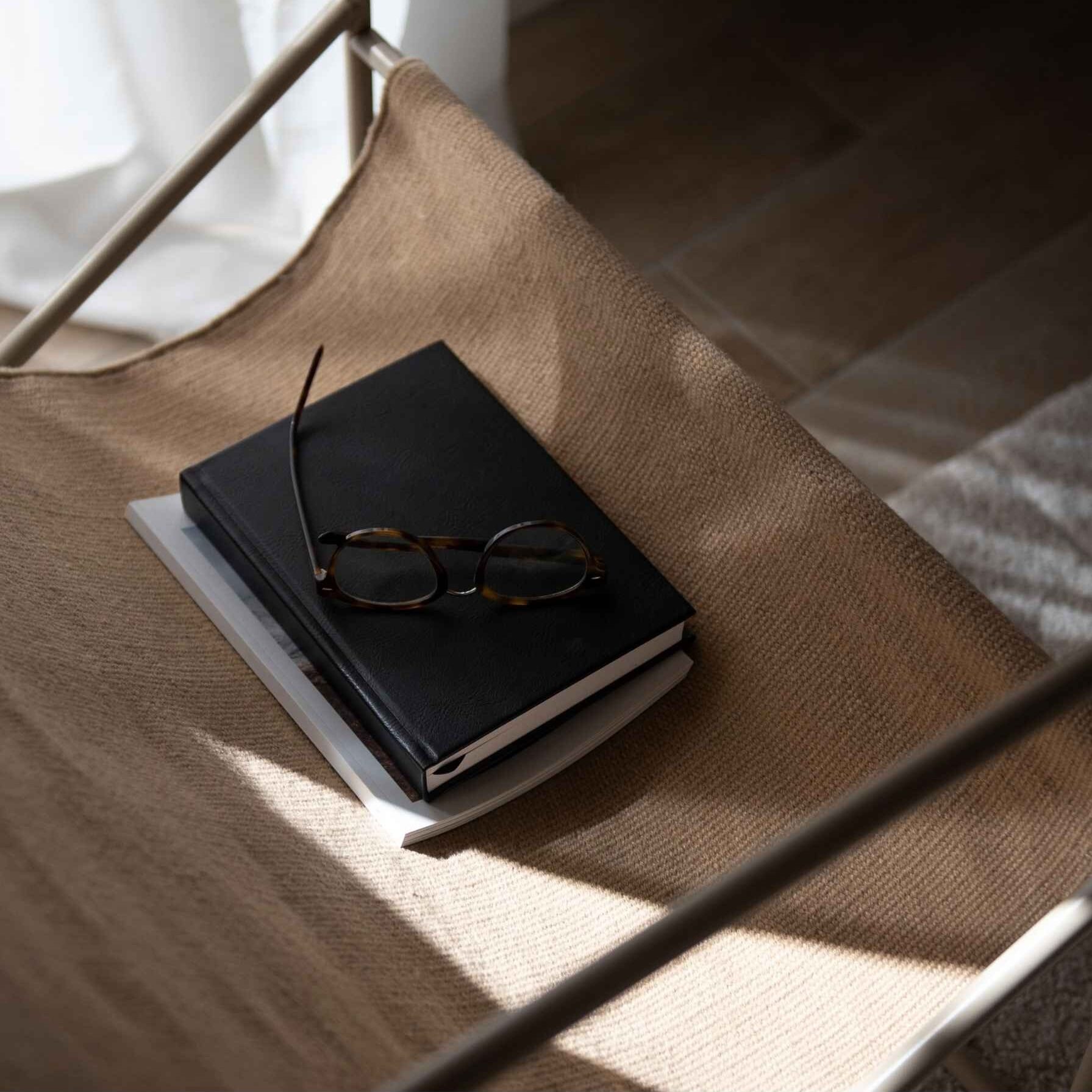 Tortoiseshell glasses resting on a black journal in a woven sling chair, lit by soft window light
