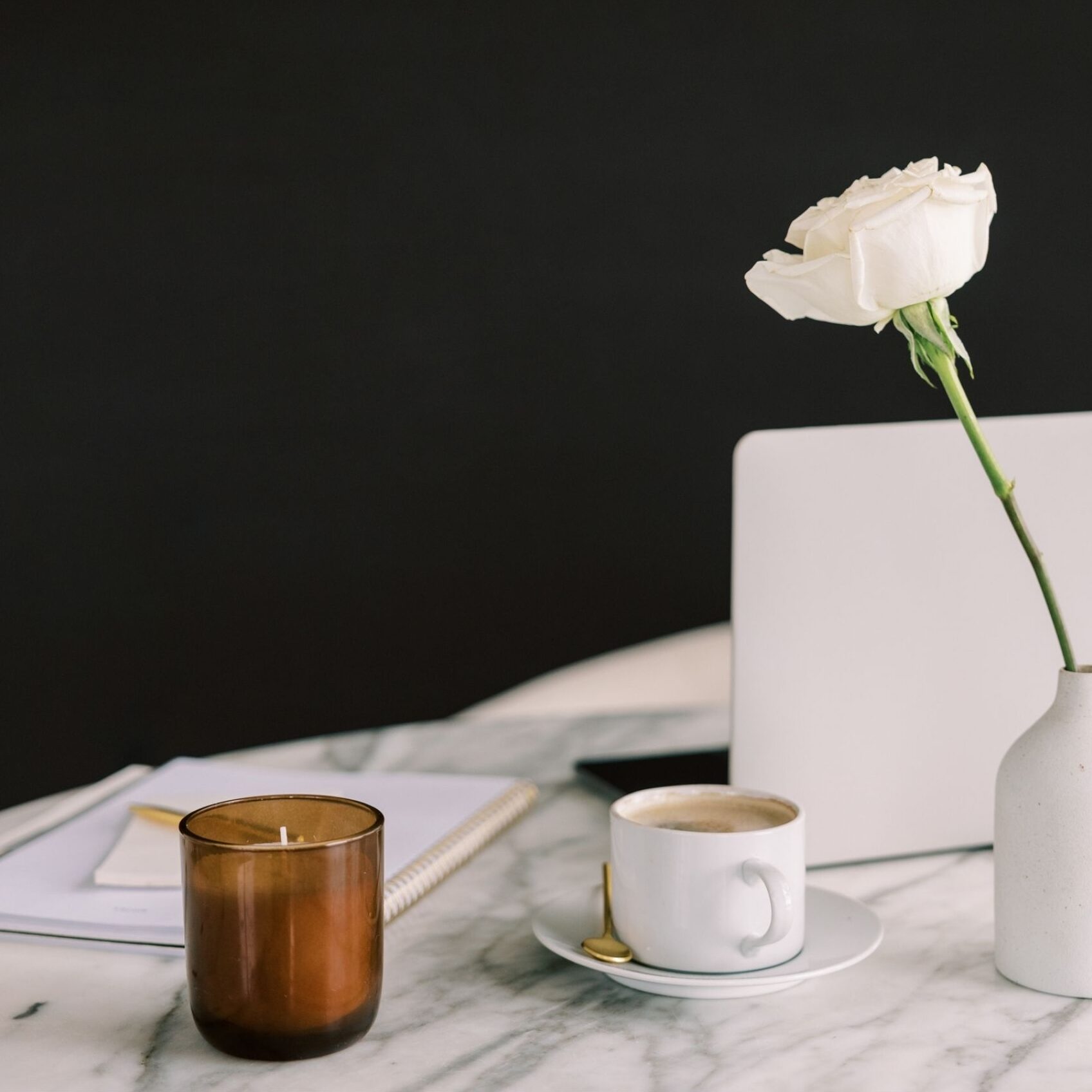 A white rose in a vase next to a laptop and coffee cup on a marble desk against a black background